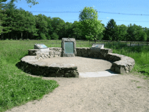Image of a stone plaque surrounded by stone walls in a circular shape. Green grass and trees are surrounding it.